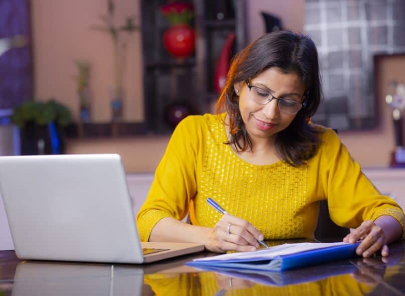 A person sits at a desk writing with a pen, a laptop to the side