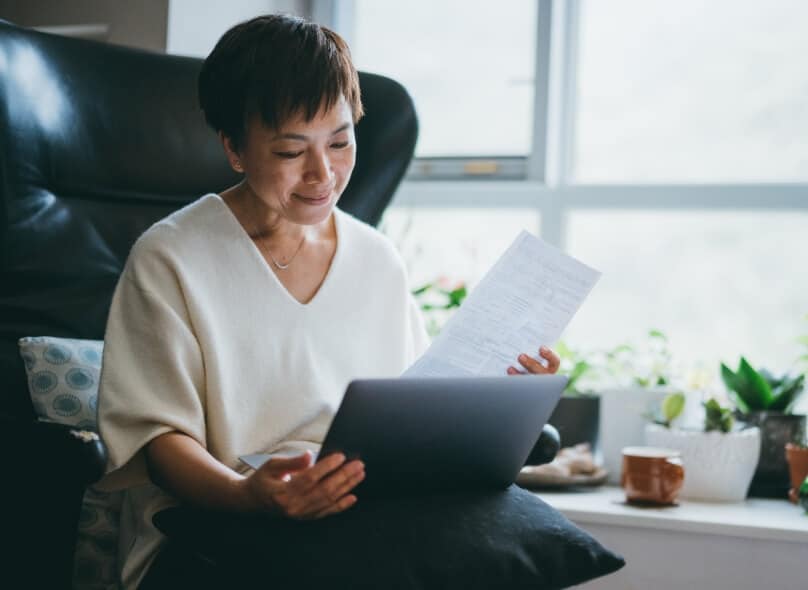 A person sits in a chair with a document in one hand and a laptop in the other