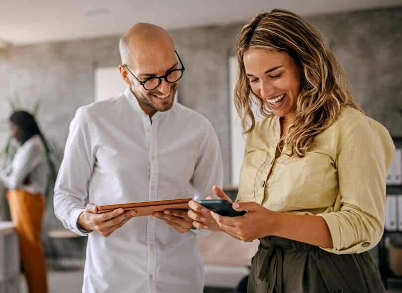 Two colleagues smile and look down at their phone and tablet respectively