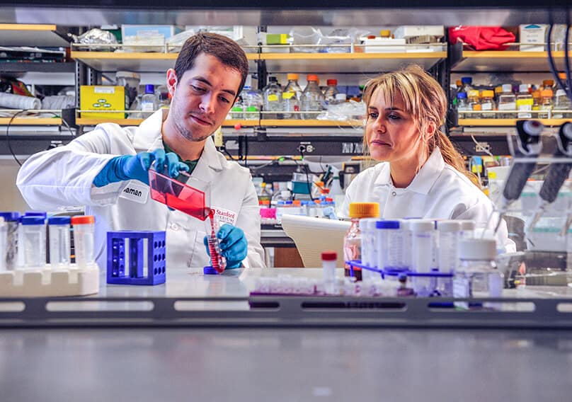 Two scientists in a lab. One scientist is pouring a red liquid out of a beaker while the other scientist looks on.